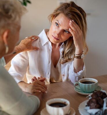 A senior mother consoling sad adult daughter indoors at home, sitting and having coffee.