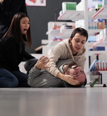 Women crying and screaming for help during person epileptic seizure in medical shop. Young man falling on floor in drugstore, having epilepsy disorder clonic stage symptoms